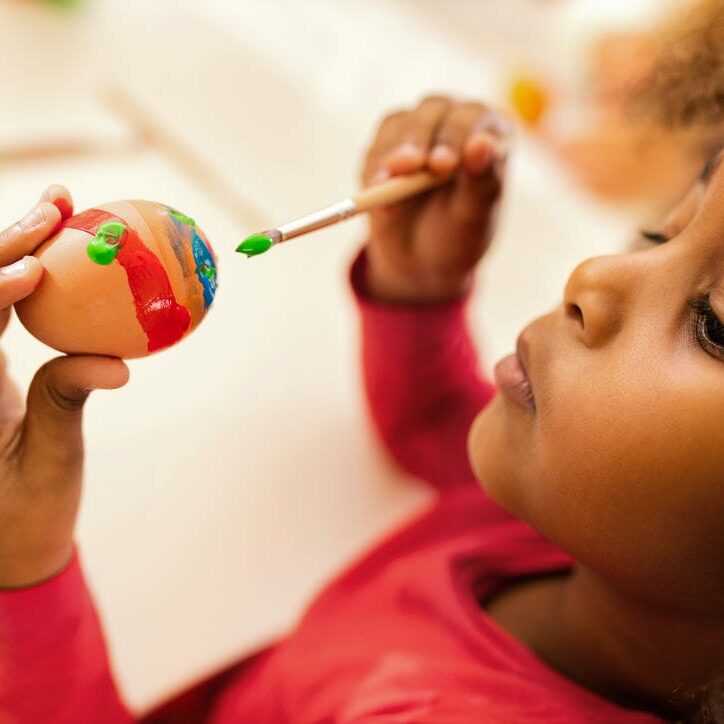 Close up of African American little girl coloring Easter egg with paintbrush.